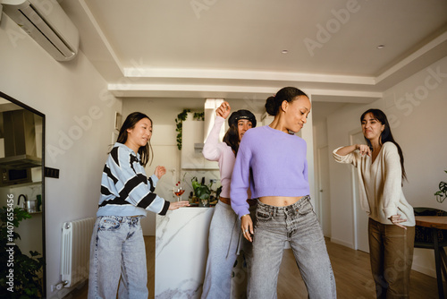 A young African American woman in her mid-20s, dancing cheerfully in her small apartment as her three female friends cheer her on while they're having a party together, wearing casual outfits