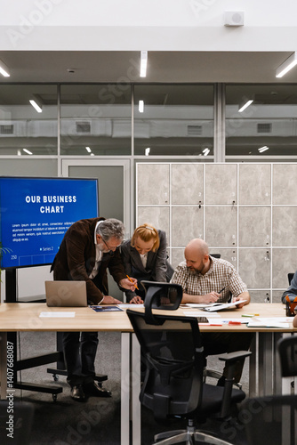 In an office space, three colleagues work at a table. An older man and a woman lean over documents, assisting a seated teammate who is writing. Concept of business discussion, working in a team.