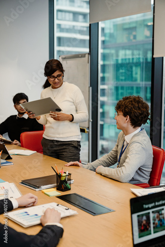 A group of interns are in a modern office. An Indian woman stands presenting a tablet while others, including a seated White man, listen and review business documents.