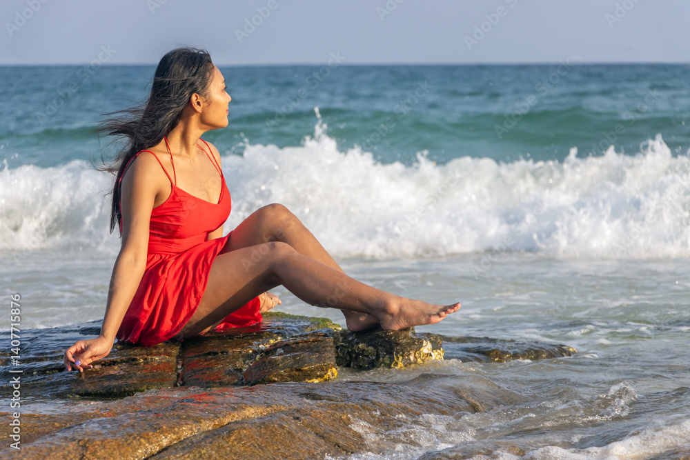 Woman in red dress sits on a rock on the coast and watches the sea waves