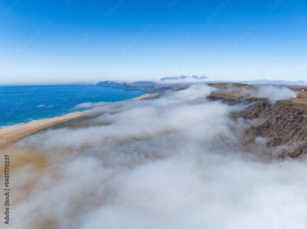 Aerial view of Rauðasandur, Iceland, where golden sands meet turquoise waters, framed by dramatic cliffs and low-hanging clouds under a radiant blue sky.