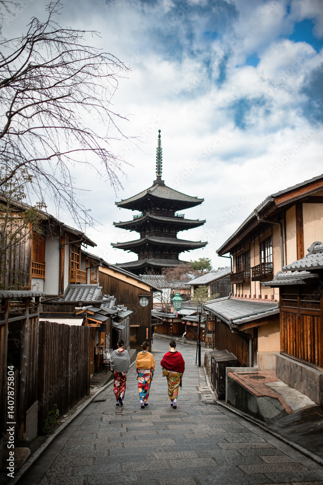 Three women in colorful kimonos walk down a traditional street in Kyoto, Japan, with the iconic Yasaka Pagoda rising majestically in the background under a partly cloudy sky.