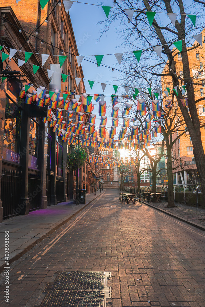 A street in Manchester, England, featuring colorful bunting, historic brick buildings, bare trees, outdoor seating, and a church like structure in the background.