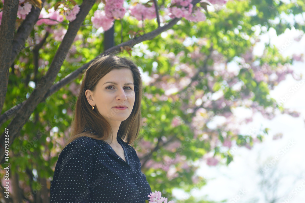 Walking in Belgrade in spring: portrait of a young smiling woman standing against the backdrop of blossoming sakura. Enjoying the beauty of nature: admiring pink flowers of Japanese cherry trees. Walking in Belgrade in spring: portrait of a young smiling woman standing against the backdrop of blossoming sakura. Enjoying the beauty of nature: admiring pink flowers of Japanese cherry trees.