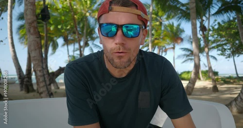 Man blogger wearing sunglasses looking at camera and speaking in mic while filming vlog in resort on tropical beach. 