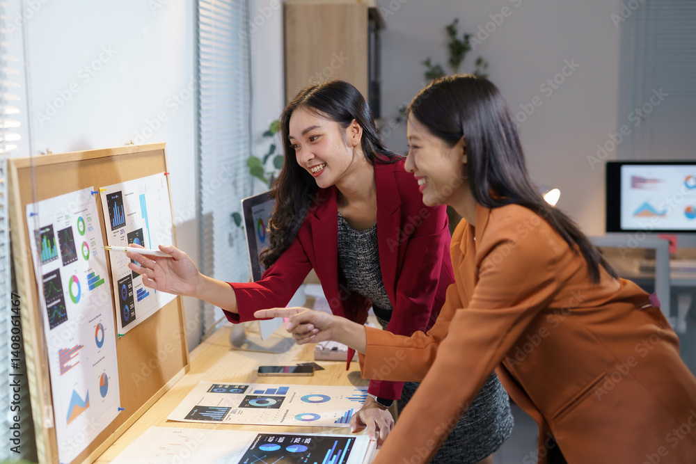 Fototapeta premium Two smiling Asian businesswomen analyzing financial charts and discussing data in modern office