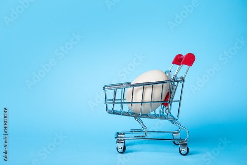 Fotografie A single white egg placed in a small shopping cart against a vibrant blue background, symbolising shopping, simplicity, and minimalism