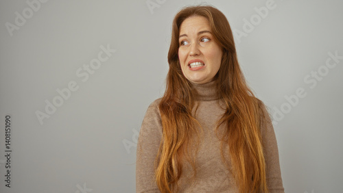 Young woman with red hair makes a face while standing in front of an isolated white background wall showcasing an uncertain expression in a casual setting.