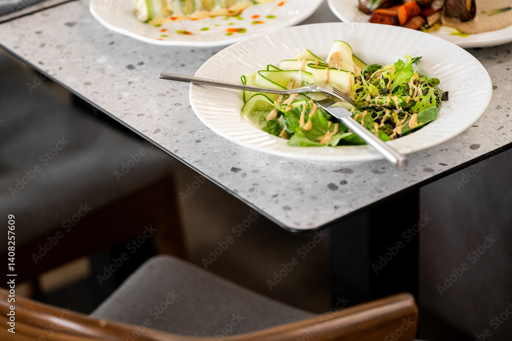 A fresh salad featuring mixed greens, sliced cucumbers, and garnishes, served on a textured plate. The dish is set elegantly on a modern table, perfect for a healthy dining experience.