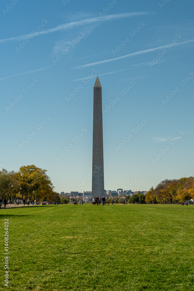 The Washington Monument rises prominently above the National Mall in Washington DC, framed by green grass, autumn trees, and a clear blue sky on a crisp fall day.