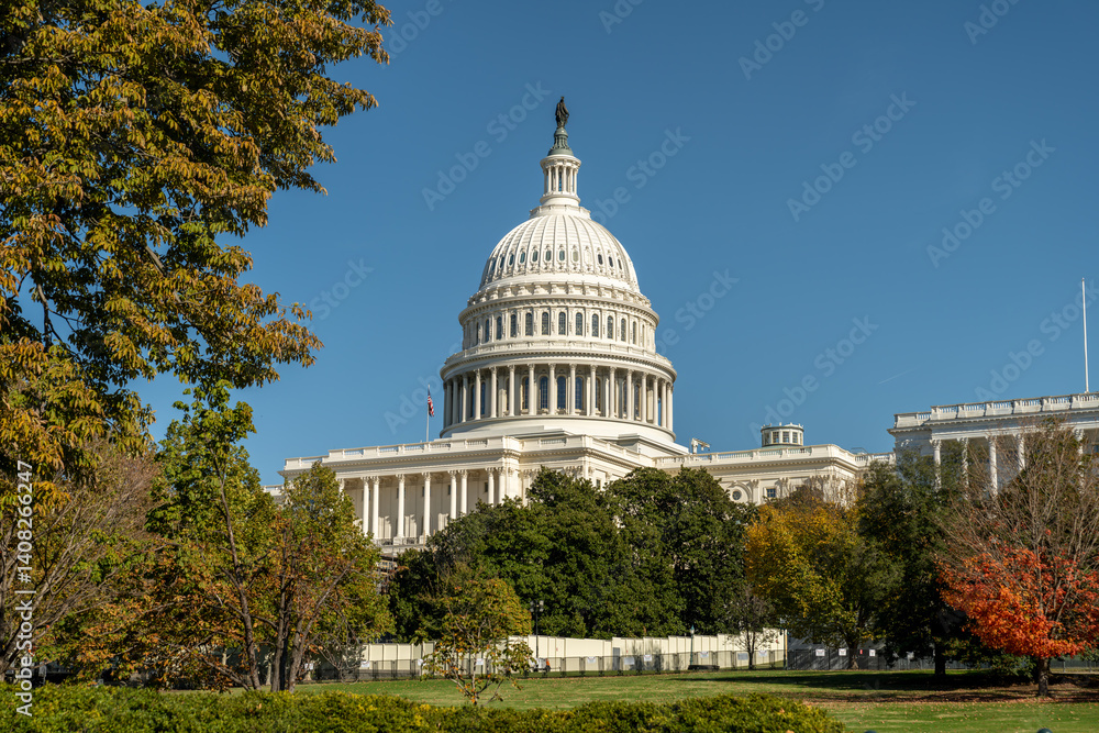 Naklejka premium The United States Capitol stands majestically on Capitol Hill, framed by vibrant autumn trees and a brilliant blue sky during peak fall in Washington DC.