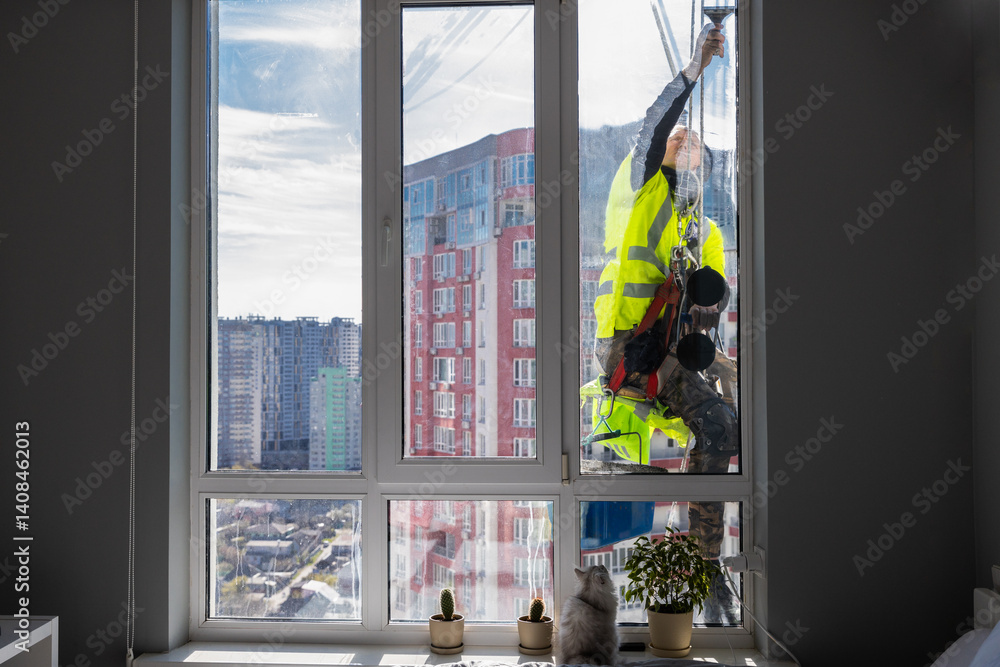 Obraz premium A dedicated window cleaner in a safety vest diligently washes large apartment windows overlooking a vibrant urban landscape while a curious cat observes from inside the cozy room.