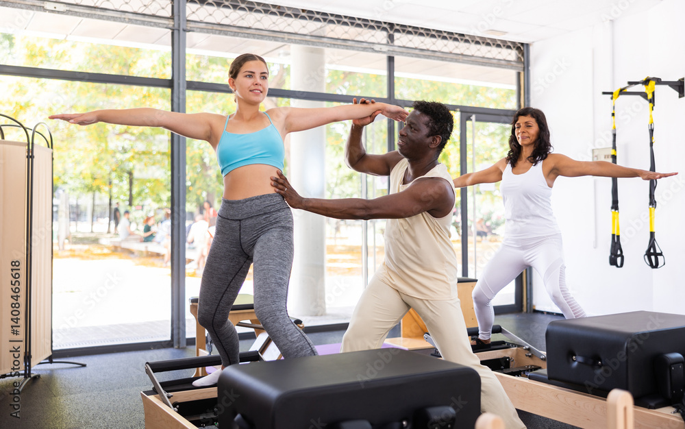 Fototapeta premium Hispanic and young european women exercising with pilates reformers. African-american man instructor assisting them with workout.