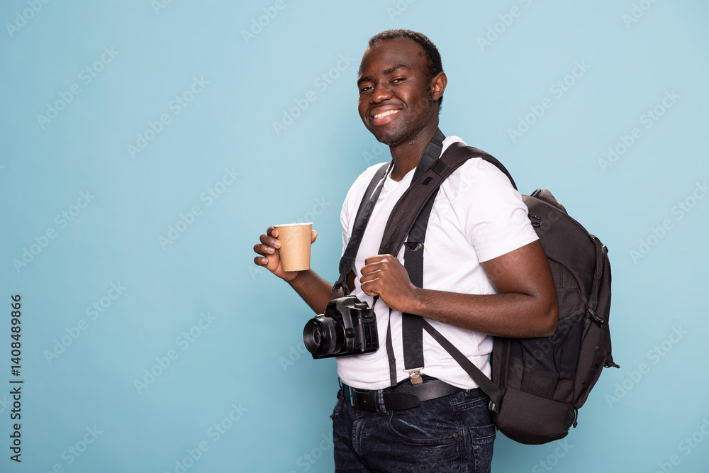 Excited male photographer stands against blue background, holding coffee cup with camera around his neck, ready for a client photoshoot. Black man with his photography gear looks optimistic in studio.
