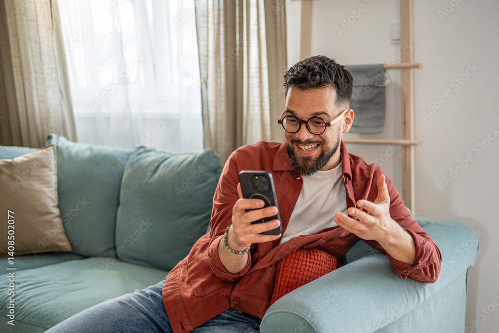 Obraz premium Happy man video calling with smartphone at home on sofa
