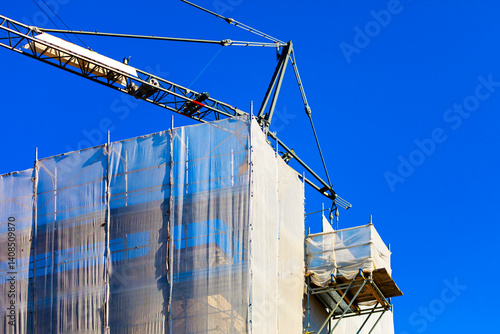 Obraz na płótnie Tall building under construction is wrapped in white scaffolding and protective netting, with a crane looming overhead against a clear blue sky. Scene captures the essence of urban development w salonie