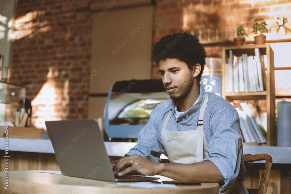 Man owner working and checking email on computer. Serious young african american guy in apron use laptop and social networks in modern loft cafe in morning after lockdown, free space Man owner working and checking email on computer. Serious young african american guy in apron use laptop and social networks in modern loft cafe in morning after lockdown, free space