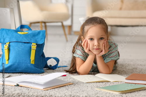 Cute little girl with notebooks and backpack lying on floor at home
