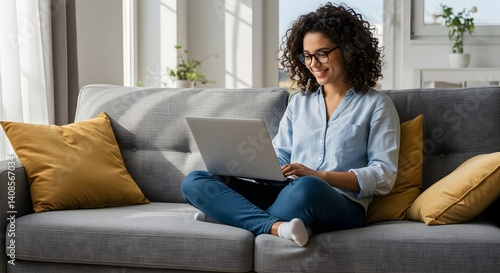 A cheerful woman with curly hair sits cross-legged on a gray couch with yellow pillows, using a laptop. Dressed casually, she enjoys the bright, cozy room filled with natural light.