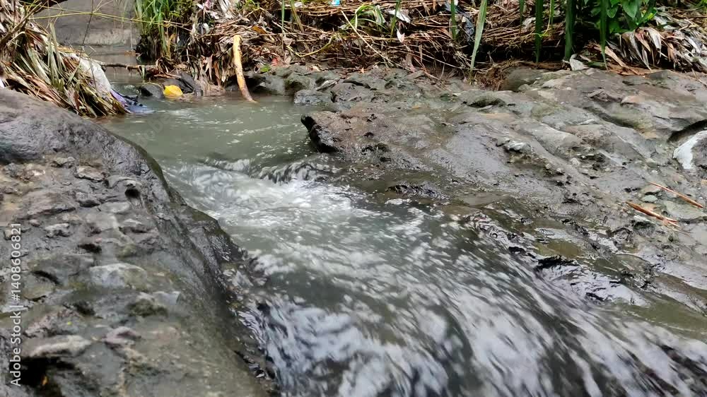 The murky stream flows over dark, uneven rocks and muddy banks with some vegetation. The murky stream flows over dark, uneven rocks and muddy banks with some vegetation.