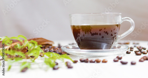 Transparent glass cup filled with steaming coffee sits on a saucer on a white table, surrounded by scattered roasted coffee beans and a small green plant, creating a cozy and inviting atmosphere