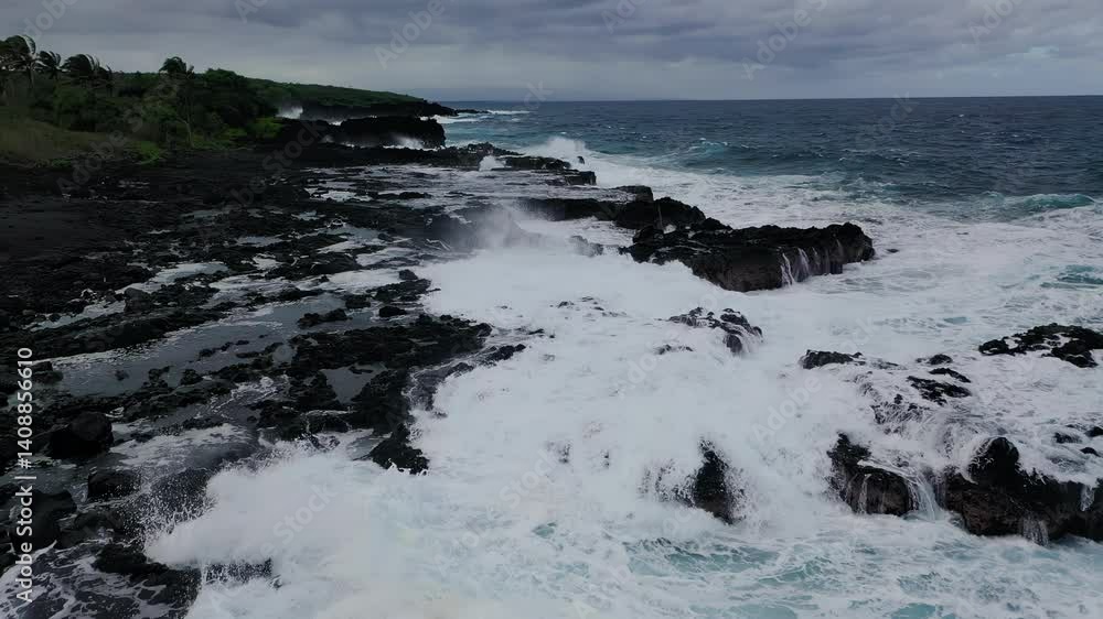 Explosive waves pound a rugged lava coastline as white water bursts upward from the black rock formations, under a cloudy tropical sky. Explosive waves pound a rugged lava coastline as white water bursts upward from the black rock formations, under a cloudy tropical sky.