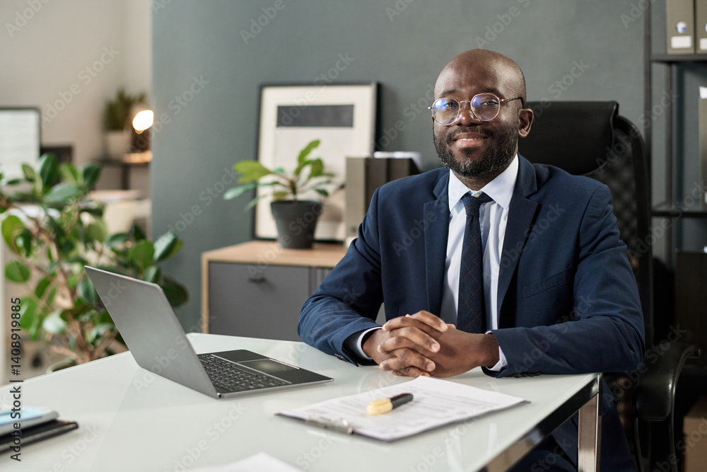 Portrait of African American manager smiling at camera while sitting at his workplace in office Portrait of African American manager smiling at camera while sitting at his workplace in office