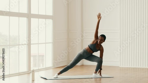 An athletic millennial woman is performing a workout indoors in her home gym. She is focused on maintaining her fitness routine while enjoying the natural light streaming in.