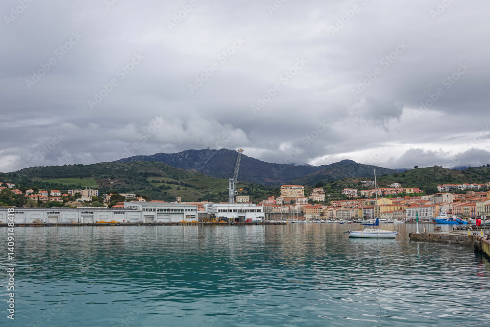 A serene harbor fills the foreground with calm waters reflecting the cloudy sky. Porte-Vendre, France