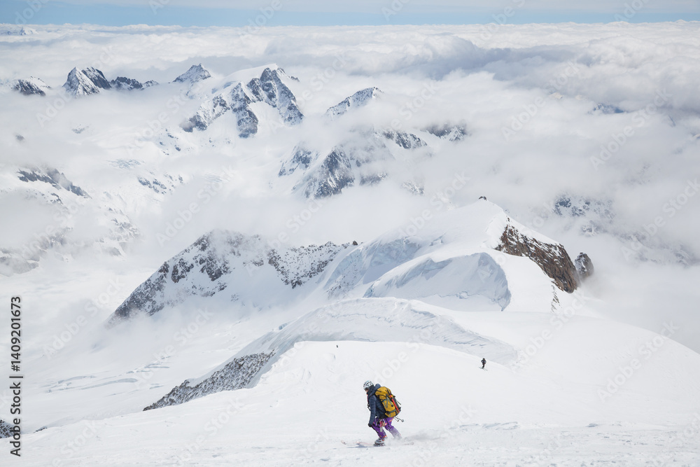 Backcountry skiers descend from mountain summit above low clouds Backcountry skiers descend from mountain summit above low clouds