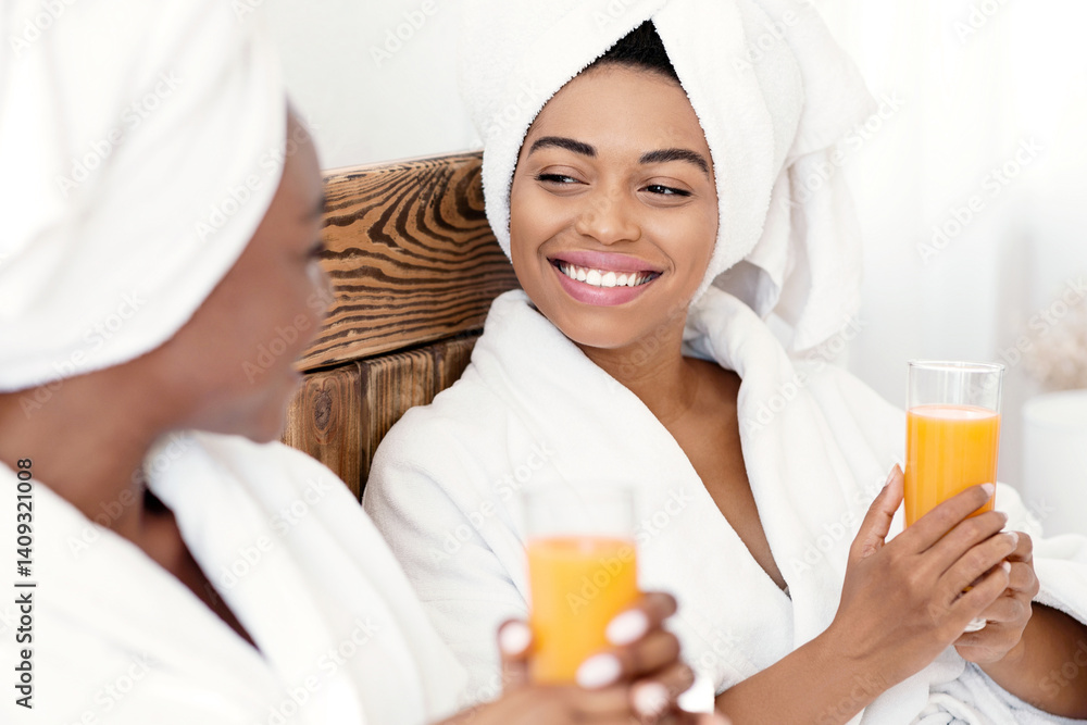 Two beautiful young black women drinking fresh orange juice together in bed at hotel. African american pretty girlfriends in bathrobes having vitamin coctail in bedroom, talking cheerfully, free space Two beautiful young black women drinking fresh orange juice together in bed at hotel. African american pretty girlfriends in bathrobes having vitamin coctail in bedroom, talking cheerfully, free space