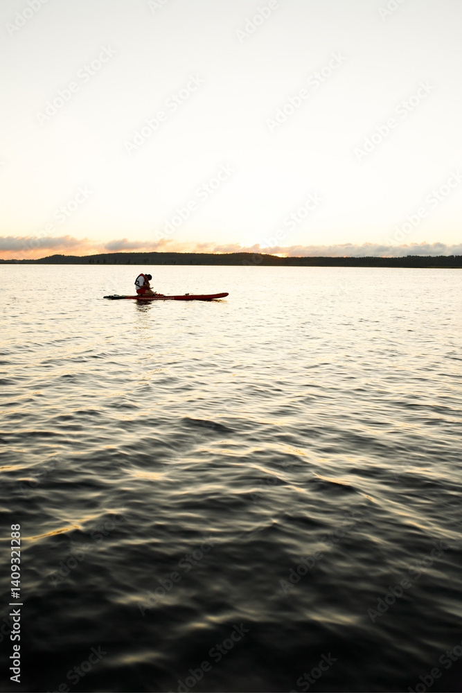 Naklejka premium Border Collie puppy in red life jacket for animals swimming on a river on a red SUP board along with his owner in a white t-shirt, red shorts and life jacket who holds a paddle on a morning dawn