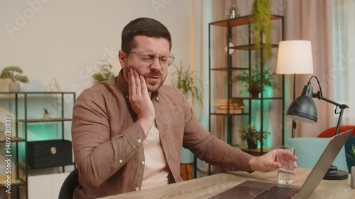 Caucasian young businessman freelancer sitting at home office table drinking cold water feeling toothache touching mouth, painful expression of dental illness, gum disease, dental disease concepts.