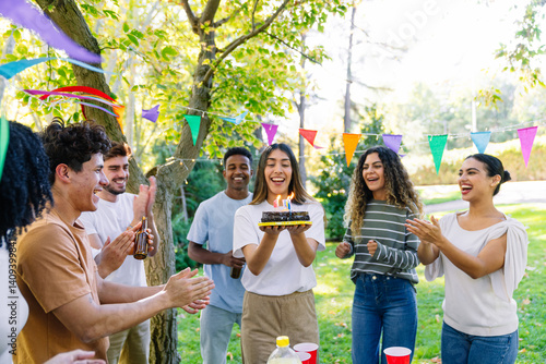 Group of cheerful multi ethnic friends clapping and celebrating birthday in a park with a birthday cake and colorful party flags