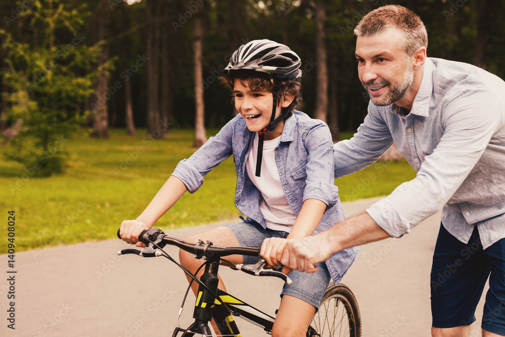Obraz premium Assistance Concept. Adult dad teaching his small son cycling at public park, pushing bike, copyspace, blurred background