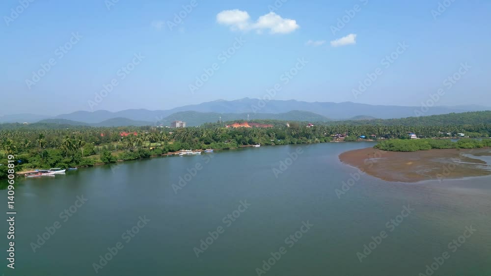 Aerial view of serene Talpona River near Rajbag Beach in Goa sparkles under the sunny sky, flanked by lush green trees and distant mountains, creating a peaceful, picturesque tropical landscape.