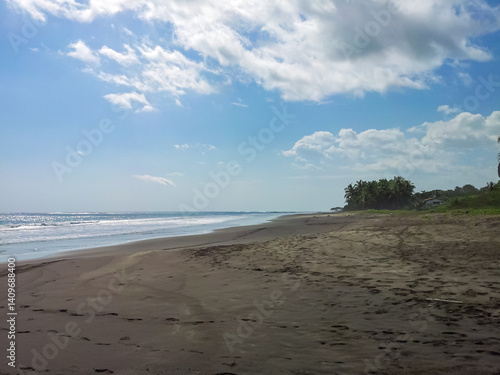 Obraz na płótnie Wide, dark sand beach, marked with footprints, extends along tropical Costa Rica coastline. Gentle ocean waves lap the shore under bright blue sky dotted with fluffy white clouds and lush vegetation w salonie