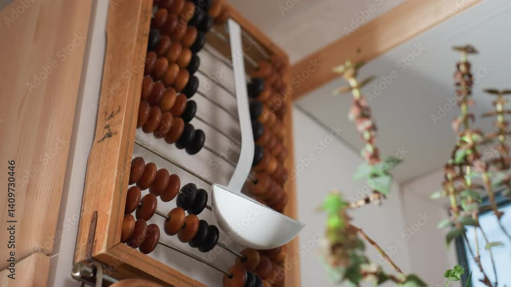 Close up of flower stems in soft focus foreground with white kitchen ladle hanging on wooden vintage abacus in bright corner near window, combining traditional decor with natural greenery