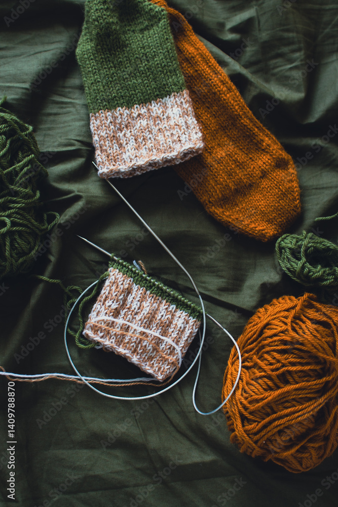 Dark green and brown yarn ball with knitted socks and knitting needles. Wool threads with deep green background. Woven crochet close up. Knitting hobby. Knitwear concept. Needlework background.