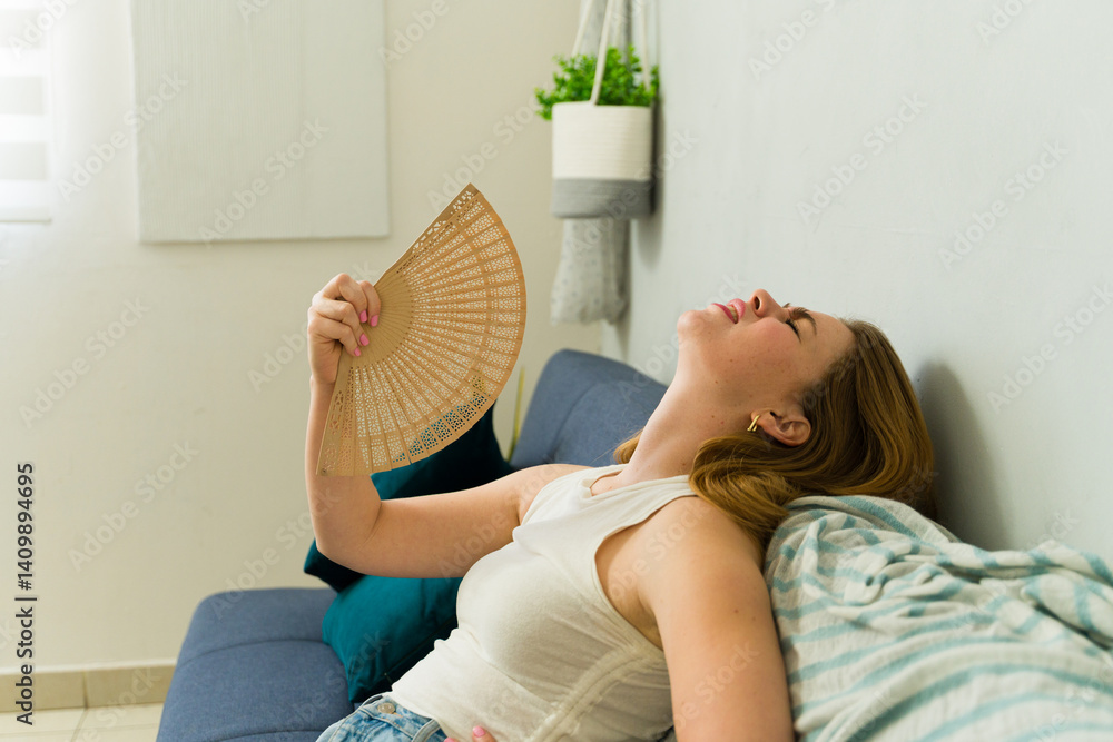 Young woman relaxing on sofa at home using hand fan during summer heat wave