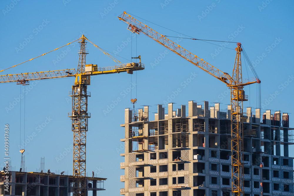 Two construction cranes working on a new residential building project, with builders constructing the concrete frame of the apartment complex against a clear blue sky Two construction cranes working on a new residential building project, with builders constructing the concrete frame of the apartment complex against a clear blue sky
