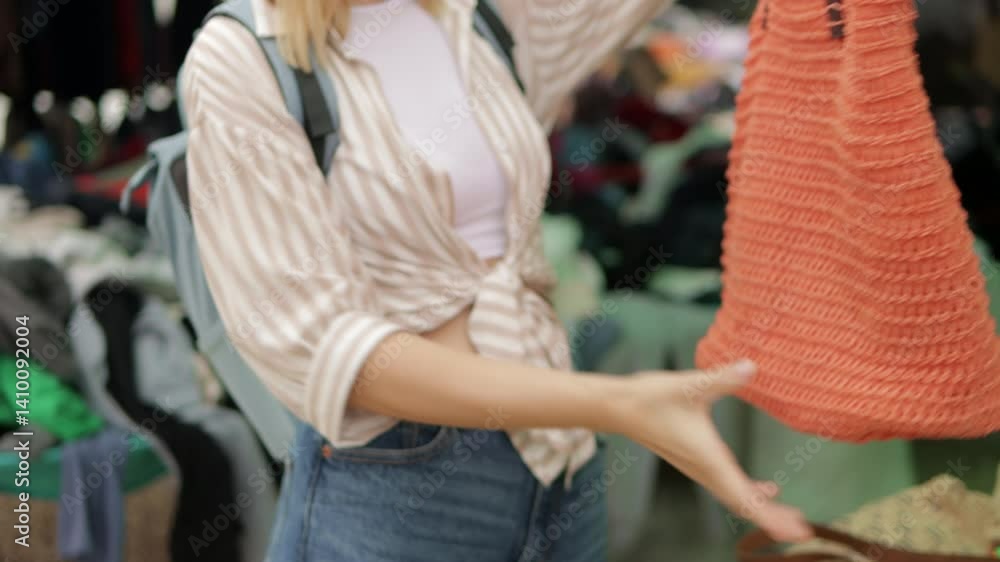 Young woman traveler carefully examining handmade knitted orange bag at vibrant outdoor street market, evaluating craftsmanship and design while exploring local artisan goods during summer vacation Young woman traveler carefully examining handmade knitted orange bag at vibrant outdoor street market, evaluating craftsmanship and design while exploring local artisan goods during summer vacation