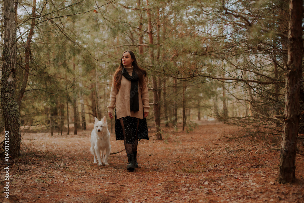 Fototapeta premium A woman in a beige sweater and black scarf walks with her white dog through a pine forest path, wearing a polka dot skirt and boots, enjoying a peaceful outdoor moment together.