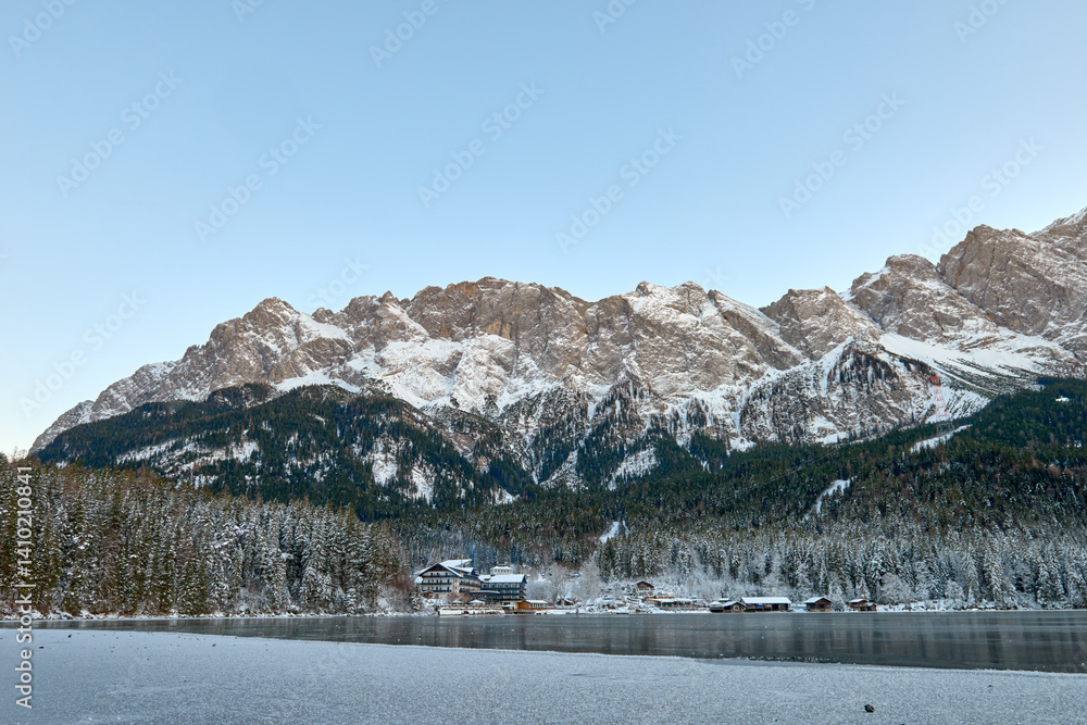 Obraz premium Twilight Alpine Wonderland: Eibsee Lake Reflects Snow-Capped Zugspitze Mountain Amidst Illuminated Pine Forest Enchanting Winter Landscape Captures Serene Beauty of German Ski Resort at Dusk.