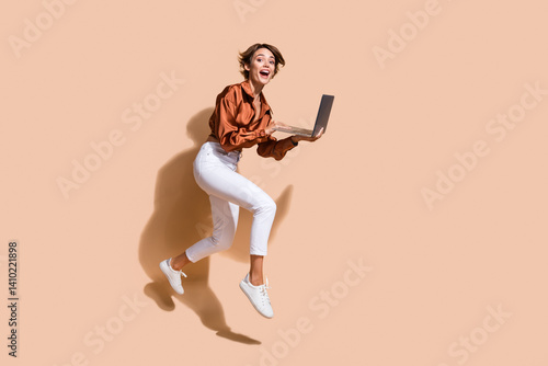 Full length photo of excited funny woman wear brown blouse jumping working modern device empty space isolated beige color background