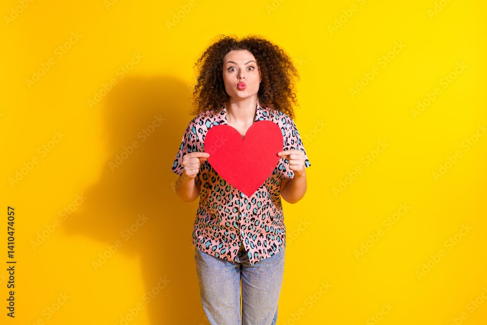Joyful woman holding a red heart shape on a vivid yellow background with charming curly hair and casual colorful shirt Joyful woman holding a red heart shape on a vivid yellow background with charming curly hair and casual colorful shirt