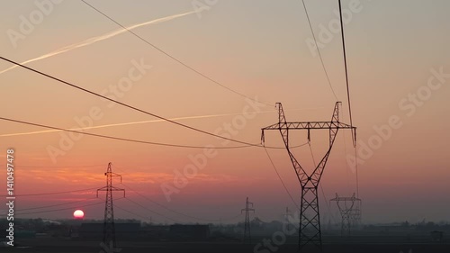 High-voltage power lines stretch across a field toward a metropolis on the horizon at sunrise, energy supply, infrastructure, and pricing, captured from an aerial, high-angle view.