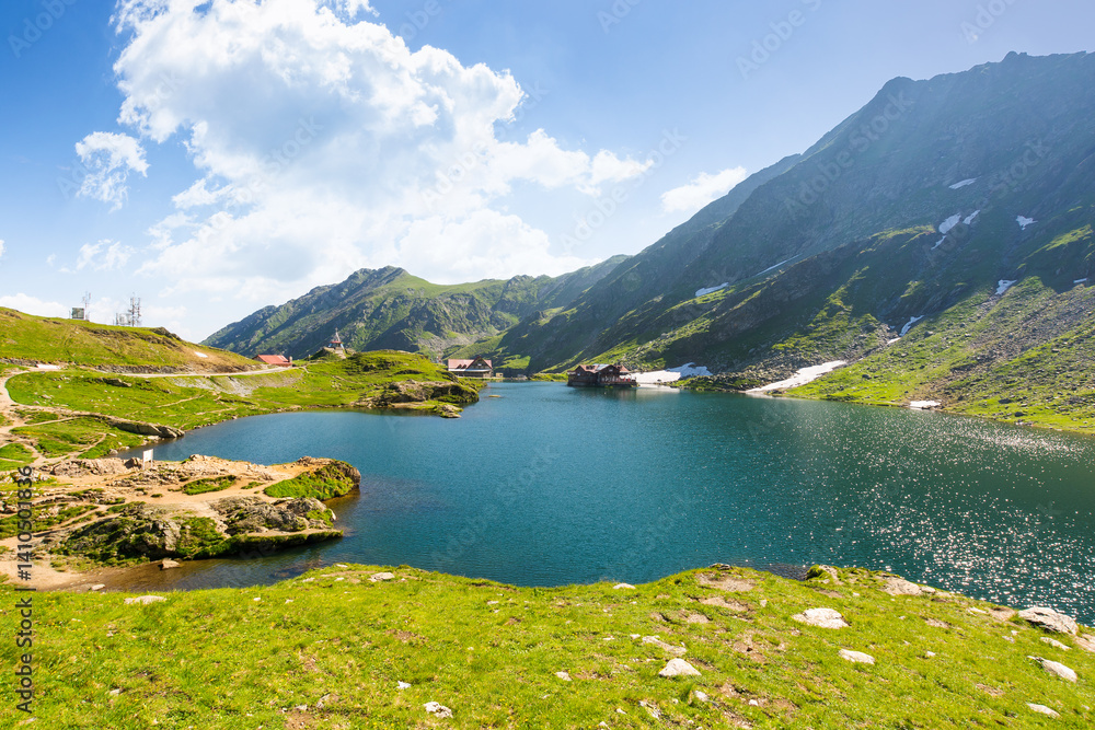 balea lake of romania. alpine landscape of fagaras mountains with pond. sunny weather with clouds on the blue sky above carpathians. snow and grass on the hillside in summer. idyllic place