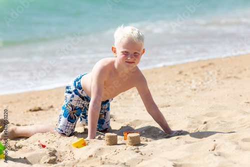 Happy child little boy playing with the sand on the beach, building sand castle, enjoying summer, playing. Advertising for tour operators, hotels, airlines. Vacation concept.