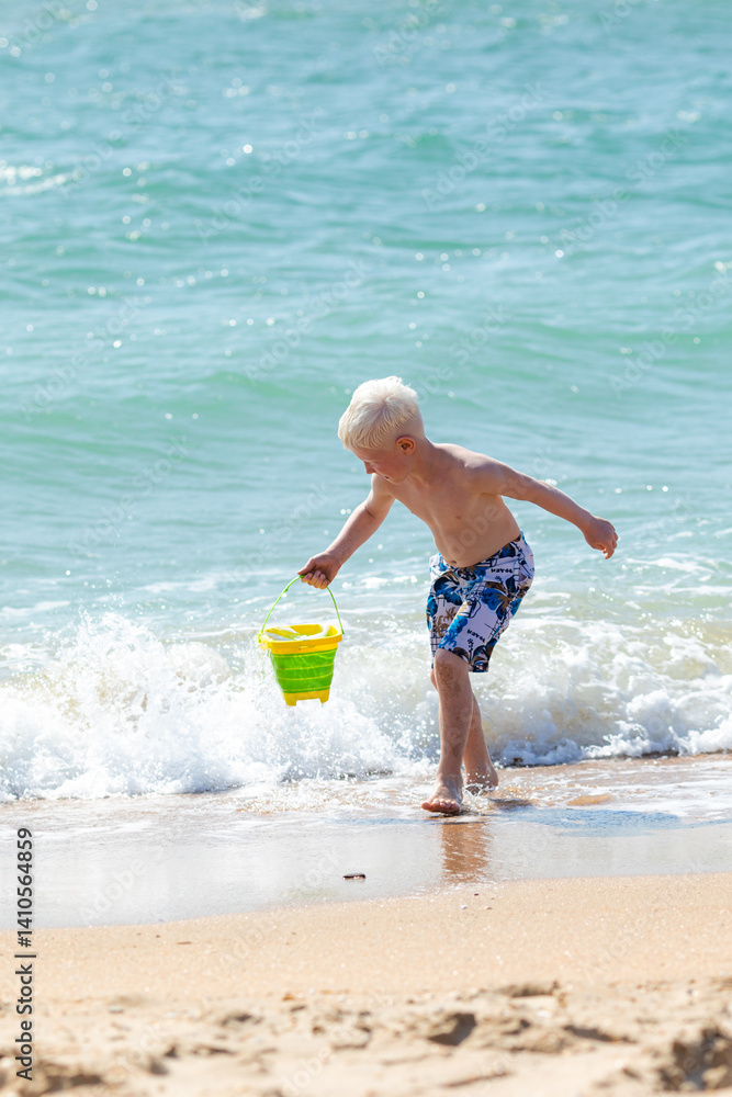 Happy child boy playing with the sand on the beach, pouring water from plastic bucket on the sea beach, building sand castle, enjoying summer. Advertising for tour operators, hotels, airlines.